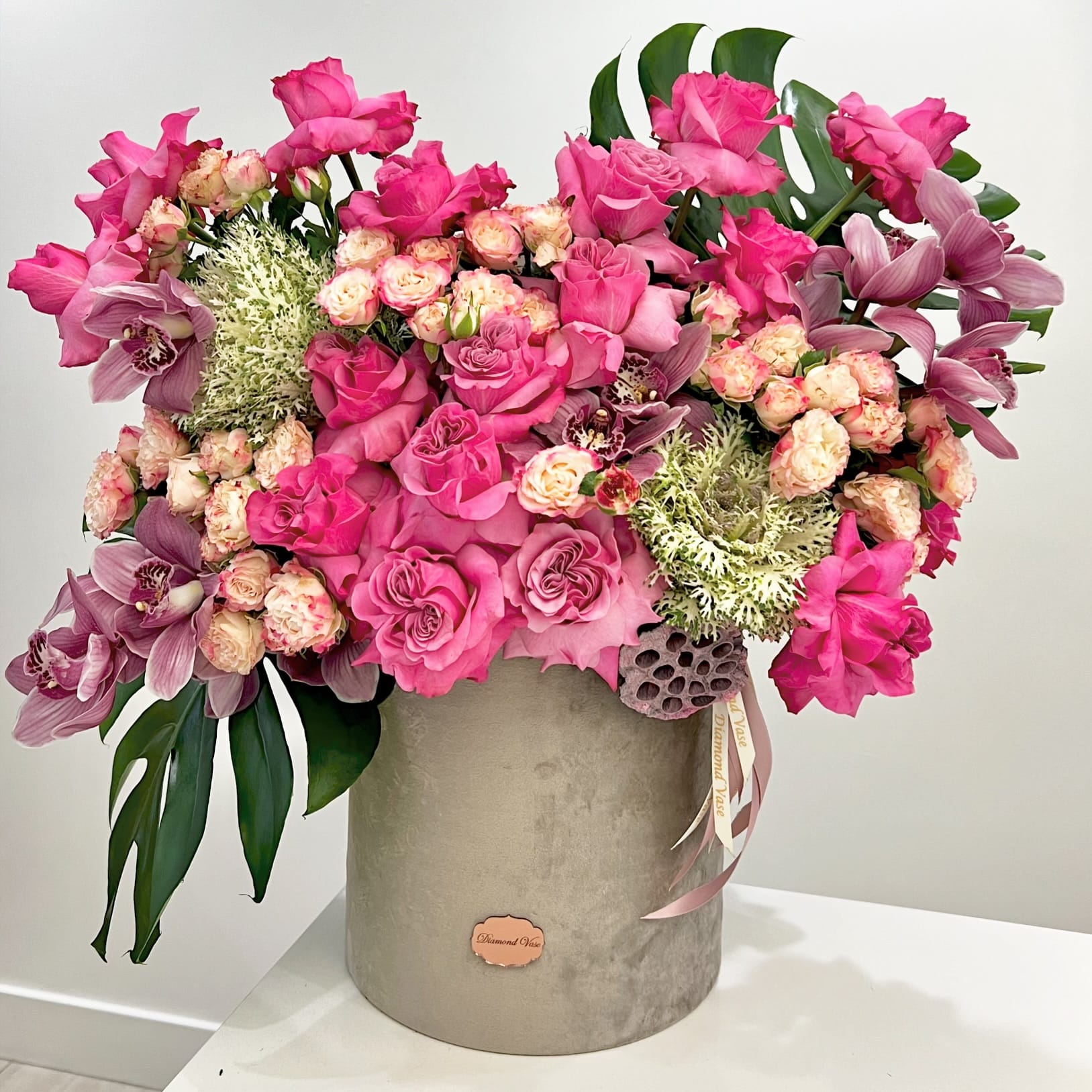 Hot-pink roses, mauve cymbidium orchids, ornamental kale, monstera and a lotus pod in a grey suede box