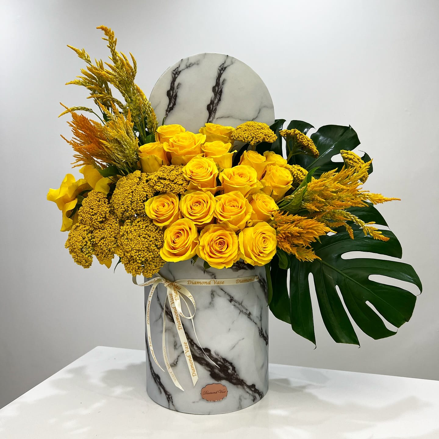 Monochrome yellow roses, yarrow and celosia with a monstera leaf in a marble-print box