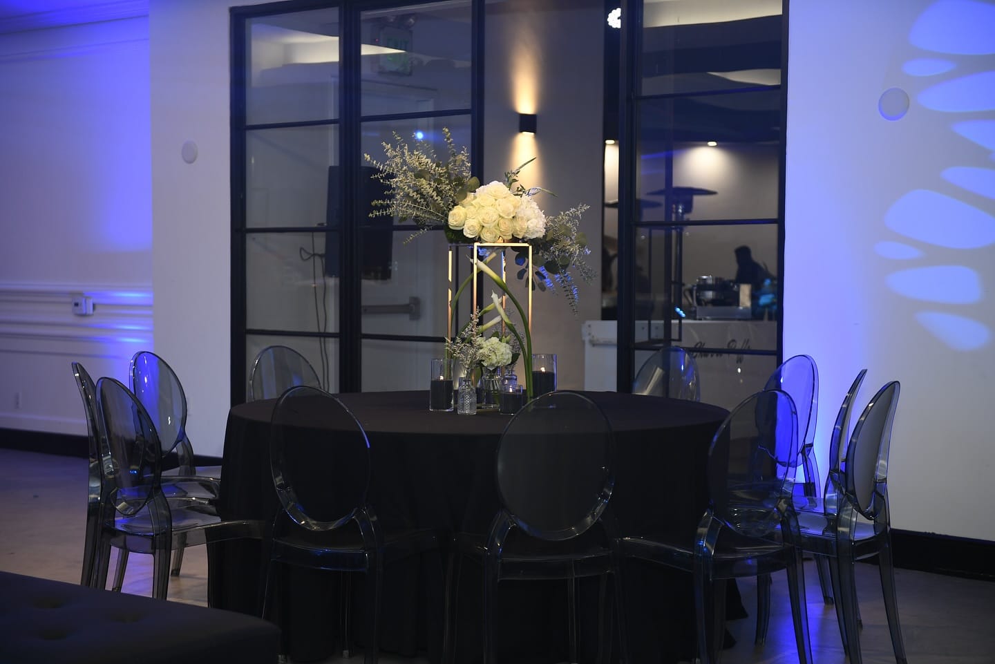 Tall gold stand of white roses, eucalyptus and calla lilies on a black-clothed round table with ghost chairs