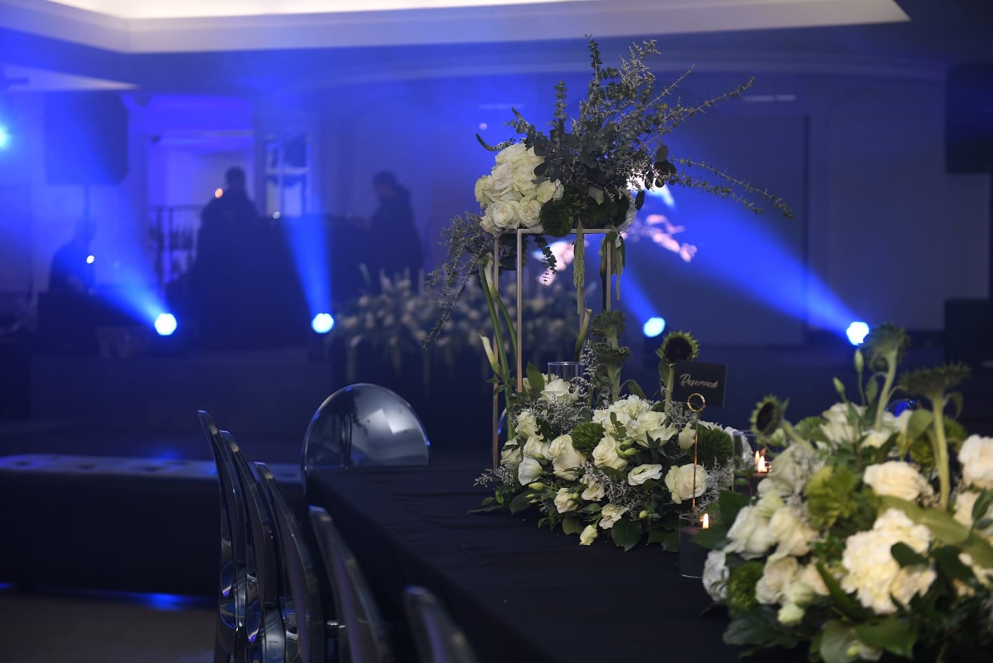 Gold stand of white roses and low centerpieces along a long black table in a hazy blue-lit ballroom