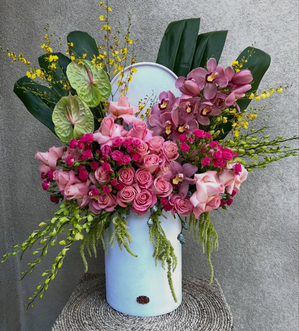 Pink roses, hot-pink spray roses, mauve cymbidium orchids, green anthuriums, yellow oncidium sprays, green dendrobium, ti leaves and trailing green amaranthus in a pale blue-white cylinder box with rounded back panel on a rattan stool against a grey stucco wall.
