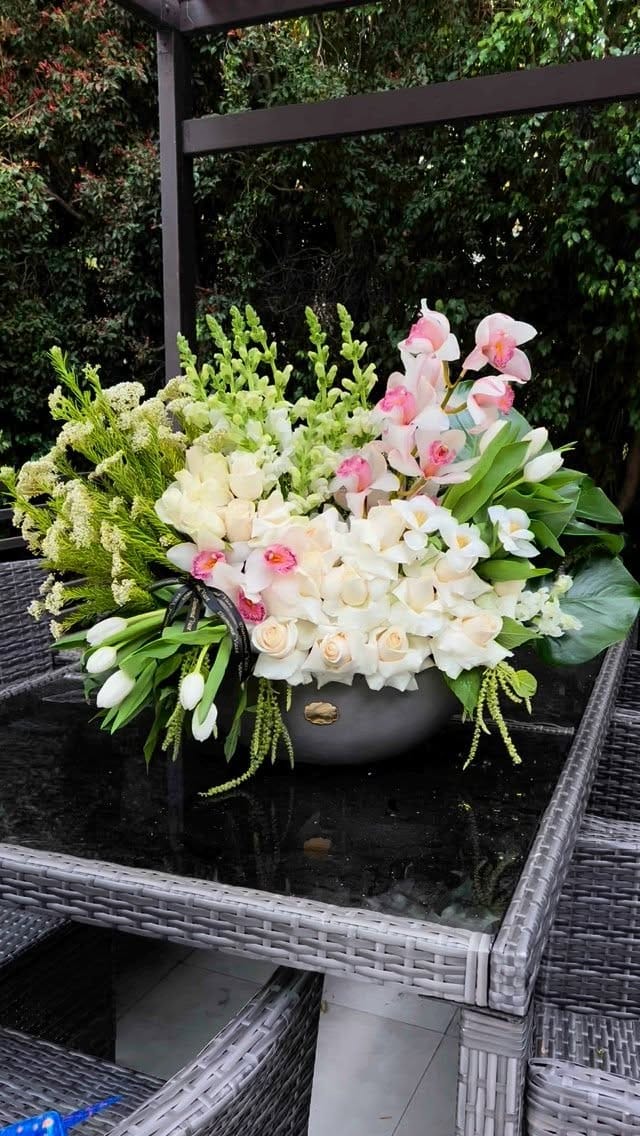 White roses, tulips and pale pink orchids in a grey concrete bowl on a patio table under a pergola