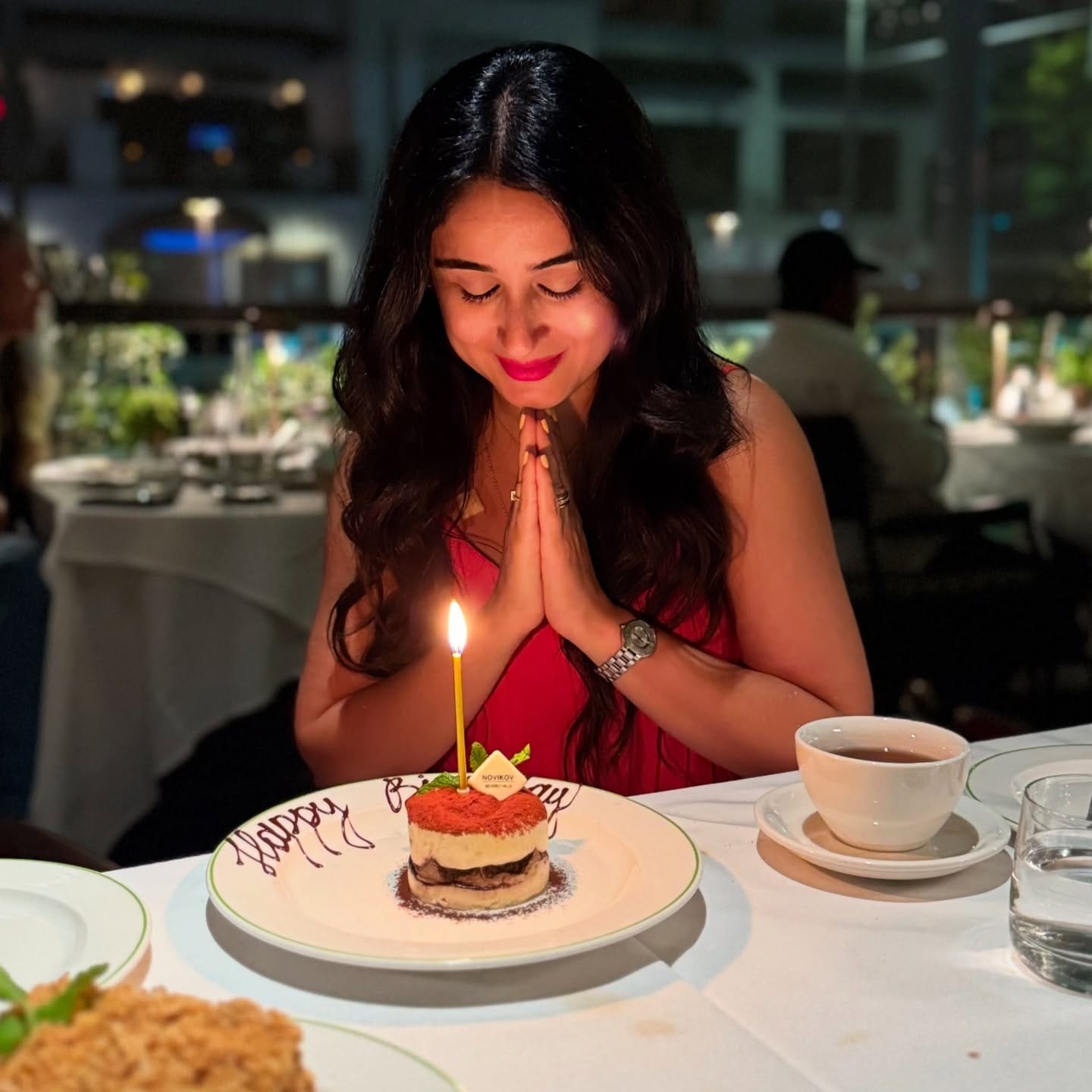 Bita in a red dress smiling at a tiramisu with a single lit candle at a restaurant table at night