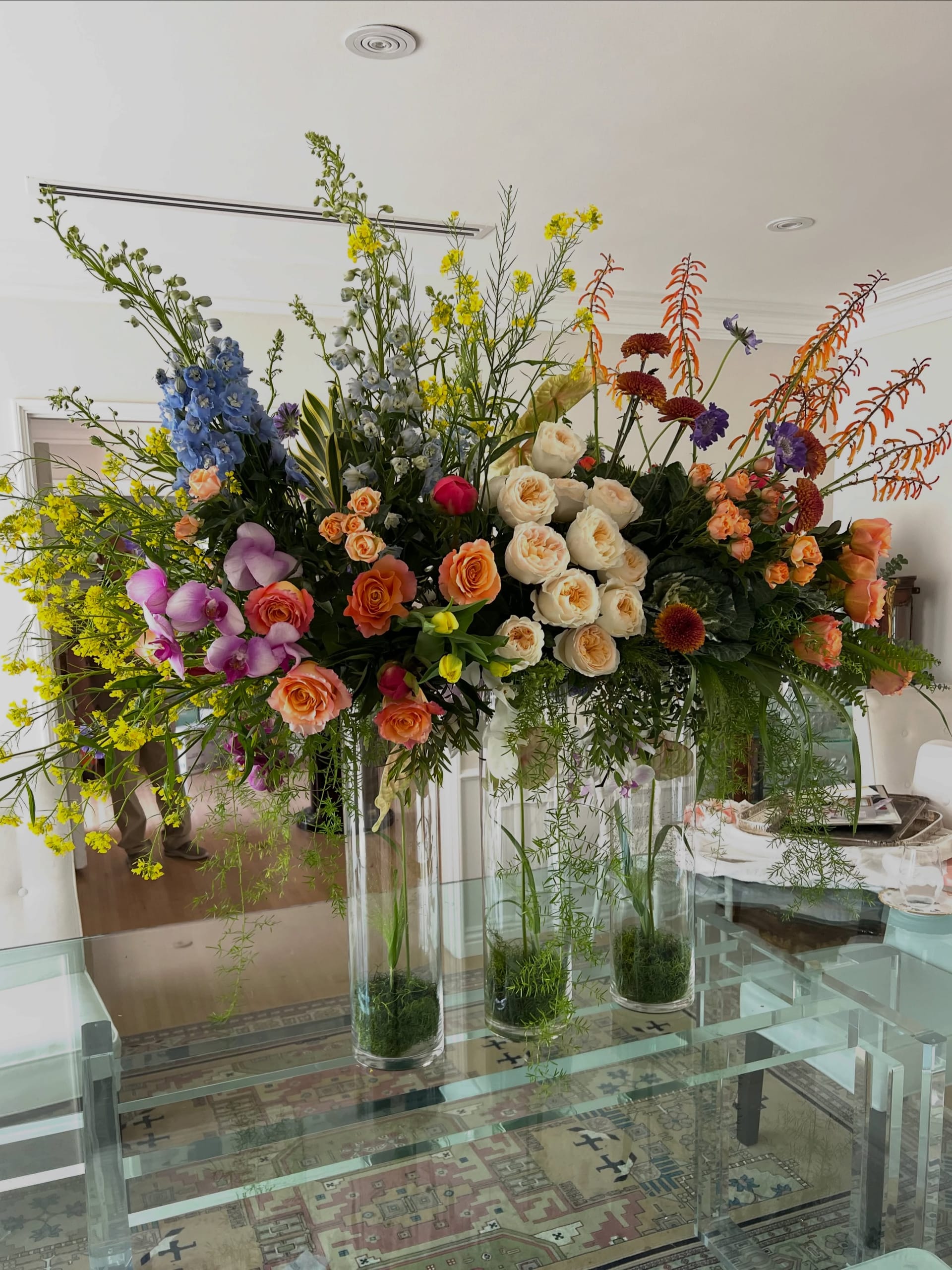 Second angle of the tall glass-cylinder arrangement with yellow mustard, blue delphinium, pink phalaenopsis, orange and cream garden roses, rust chrysanthemums and ornamental kale on a lucite dining table.