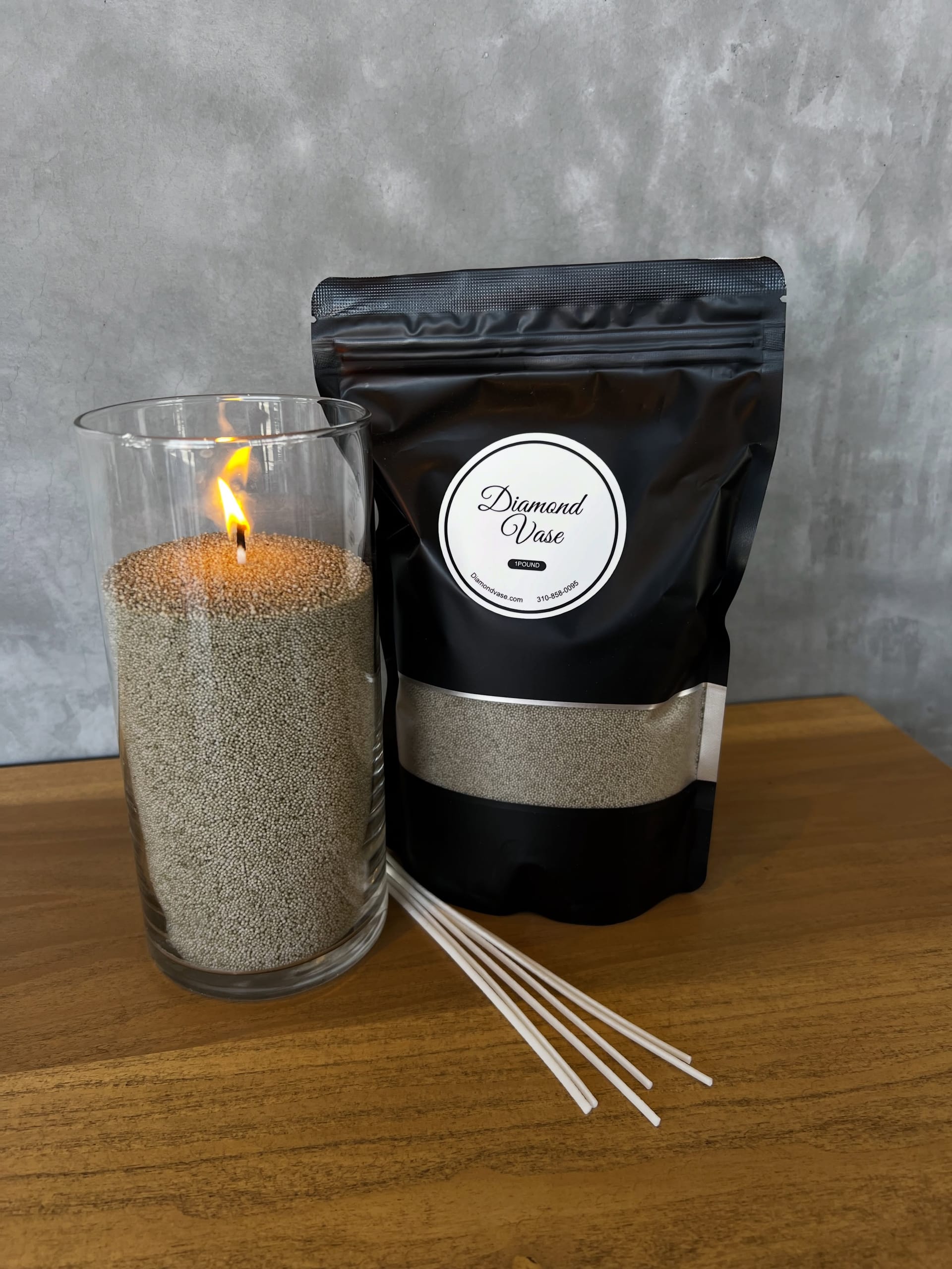 A silver-grey Diamond Vase sand candle burning in a glass cylinder beside its black pouch