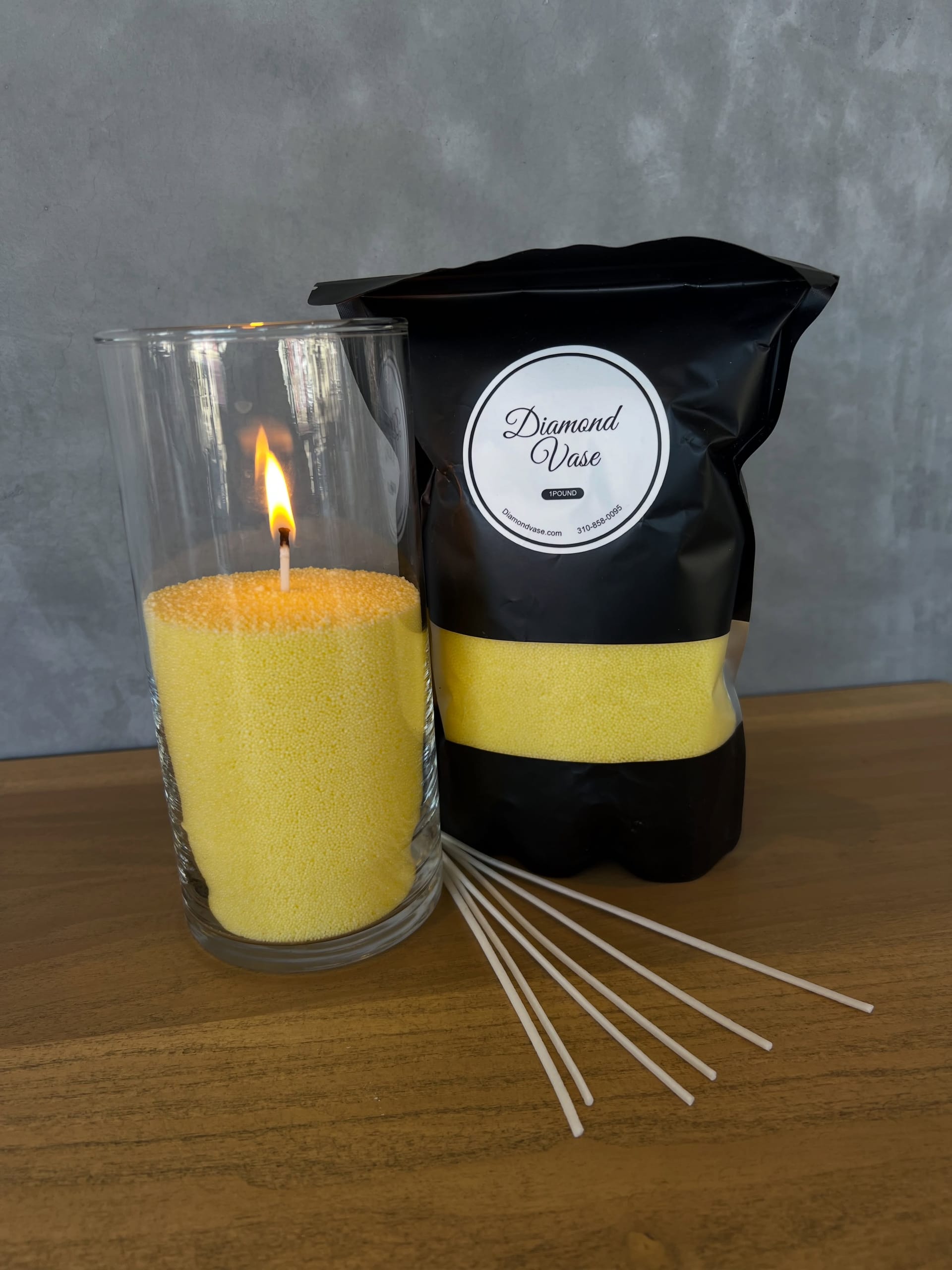 Pale yellow sand candle burning in a clear glass cylinder beside its black pouch on a wood table