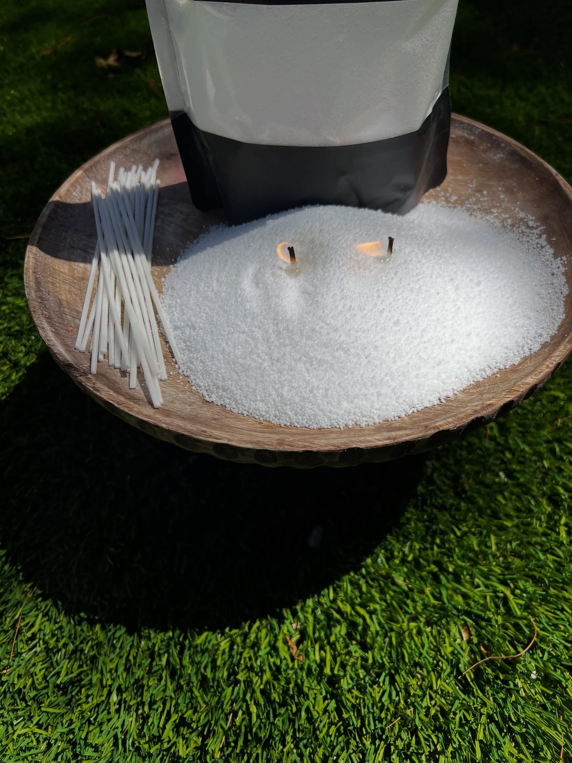 White sand candle wax with two lit wicks heaped on a wooden pedestal bowl on artificial grass, a bundle of wicks beside it and the pouch cropped at the top.