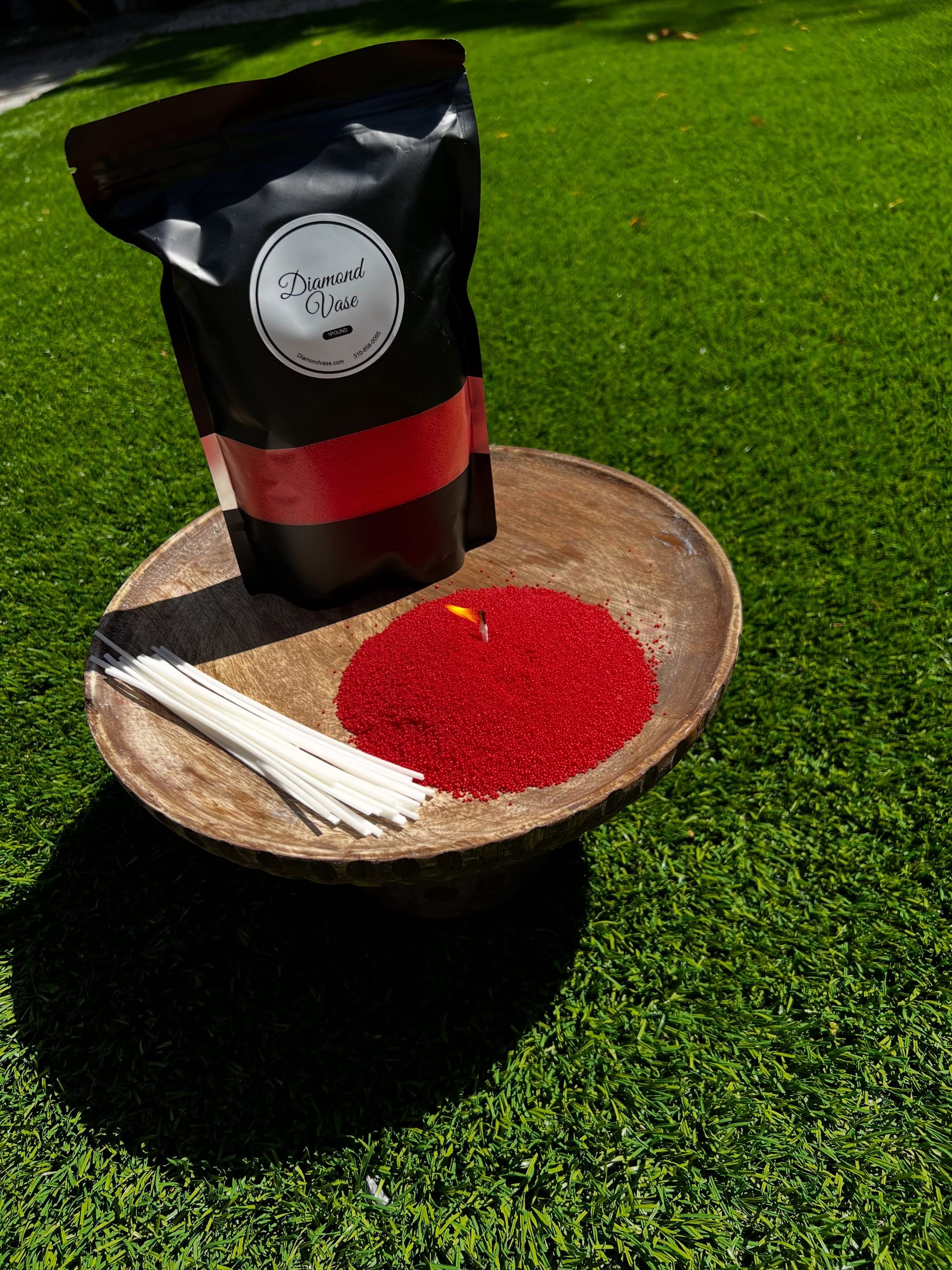 Bright red sand candle wax with a lit wick on a wooden pedestal bowl on green grass beside a black Diamond Vase pouch with a red window and a bundle of wicks.