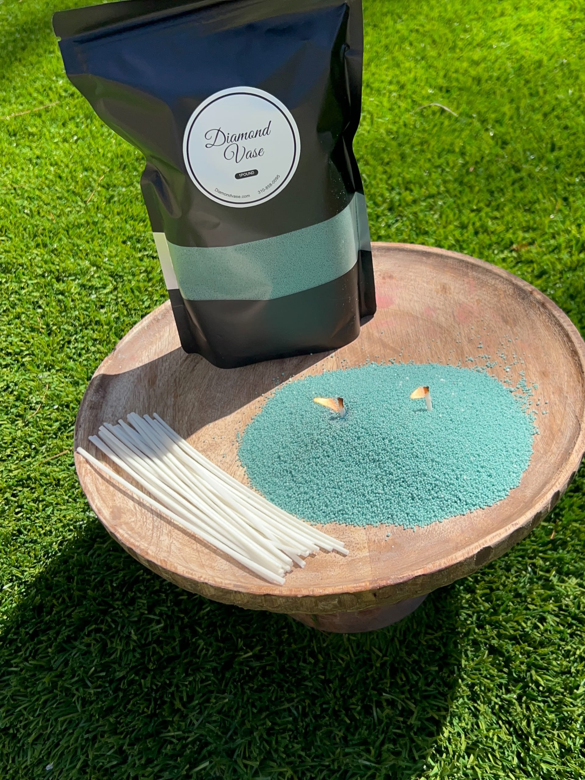 Teal-aqua sand candle wax with two lit wicks on a wooden pedestal bowl on grass beside a black Diamond Vase pouch with a teal window and white wicks.