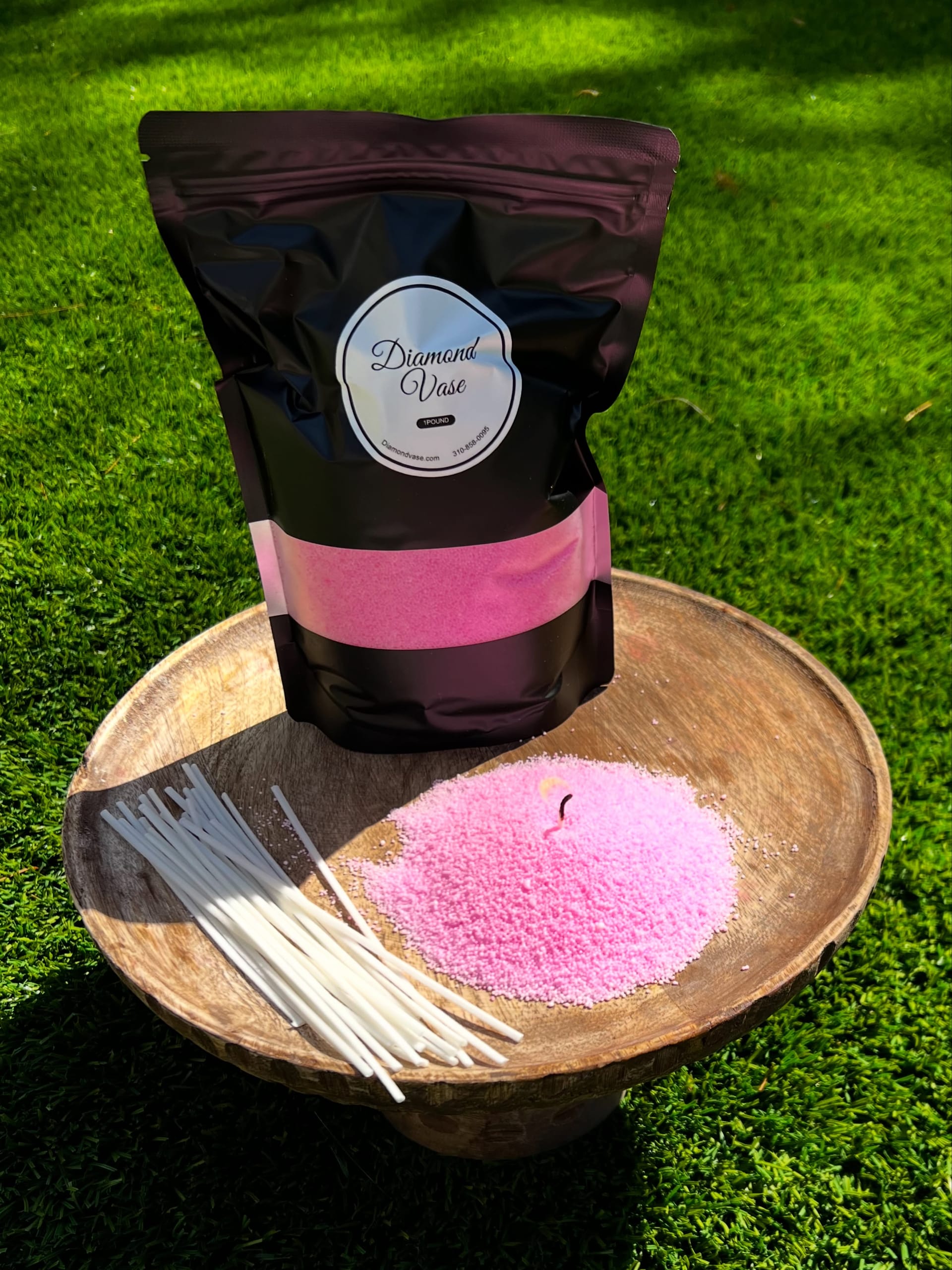 Bubblegum-pink sand candle wax with a lit wick on a wooden pedestal bowl on grass beside a black Diamond Vase pouch with a pink window and a fan of wicks.