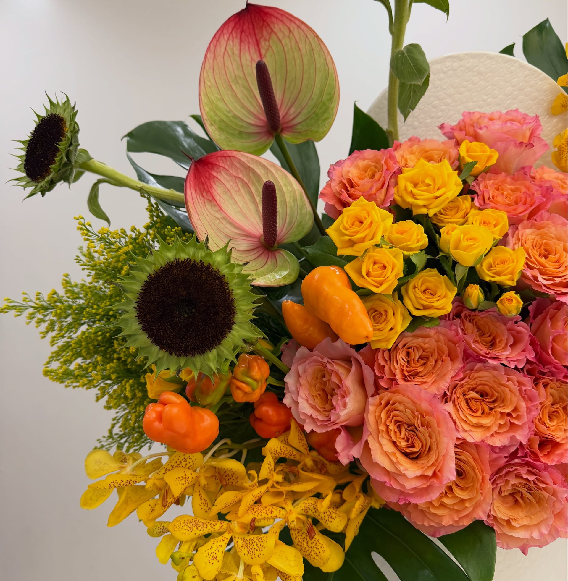 Close-up of two pink-veined green anthuriums, a dark sunflower head, orange peppers, yellow spray roses, orange garden roses, yellow mokara orchids and solidago beside the white textured box.