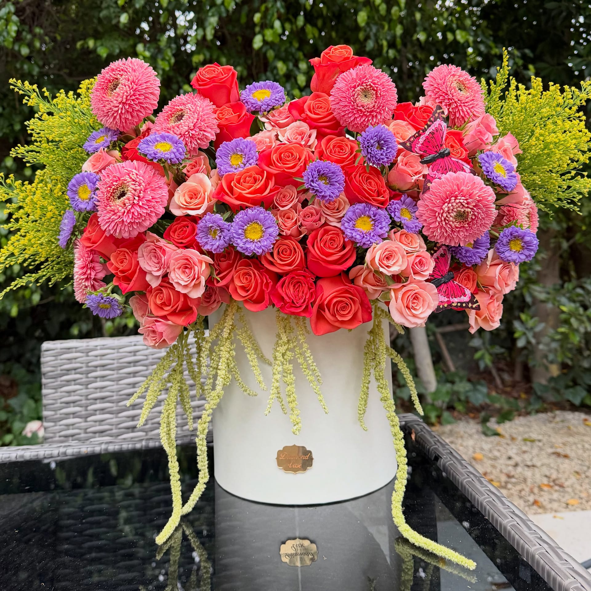 Coral garden roses, pink and purple asters and trailing green amaranthus in a white Diamond Vase box on a garden patio